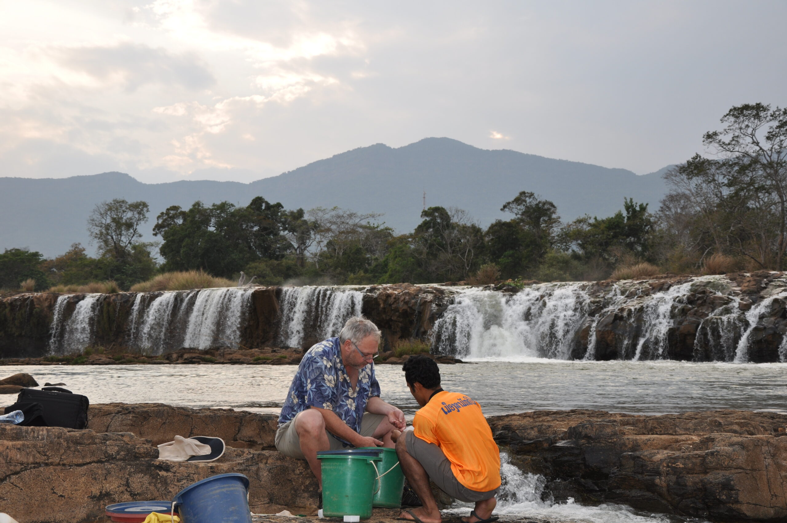 A photograph of Maurice Kottelat sitting on a rock at the edge of a lake with another man, both of them are looking in buckets.