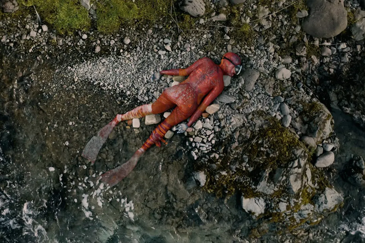 A person in a red fish costume and snorkel lying face down on rocks next to water.