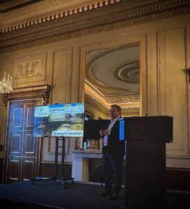 Michael Baltzer on stage talking next to a screen in a nice looking room.