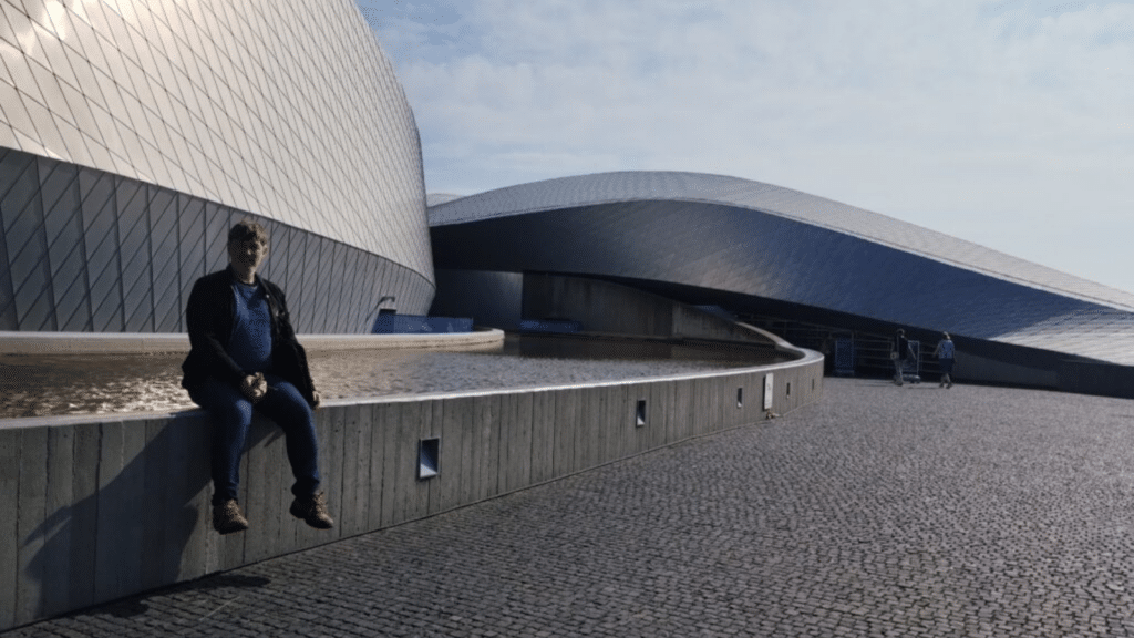 A man sits on a grey wall in front of a large and low concrete and metal building.