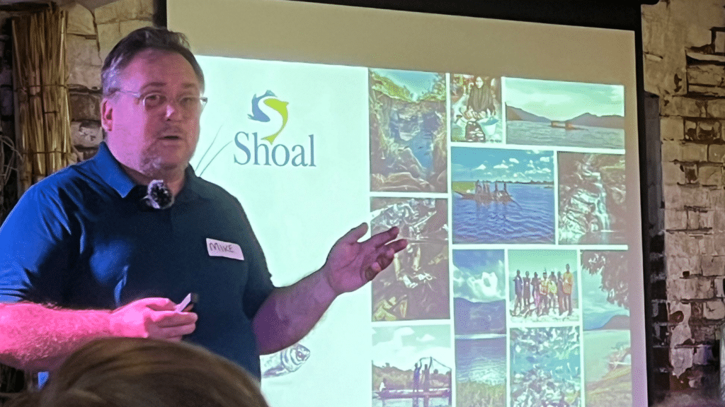 A man stands infront of projector screen, with a slide that says SHOAL next to multiple photographs of fish.