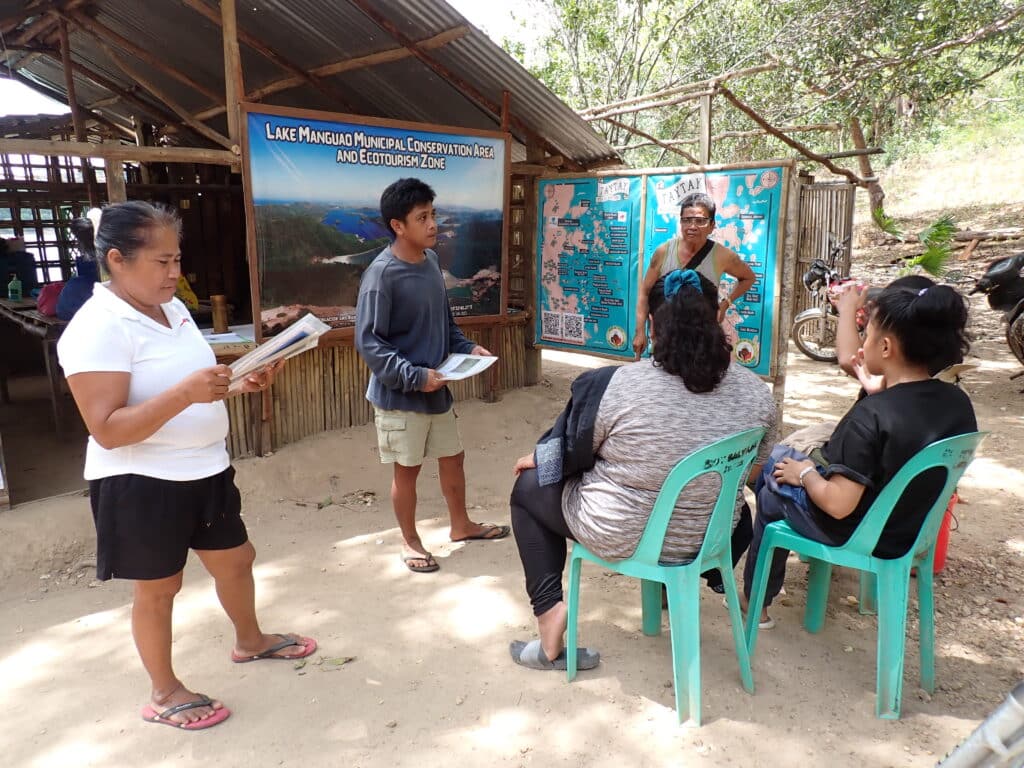 Three people sitting listening to three people presenting to them with a board behind saying: "Lake Manguao Municipal Conservation Area and Ecotourism Zone"