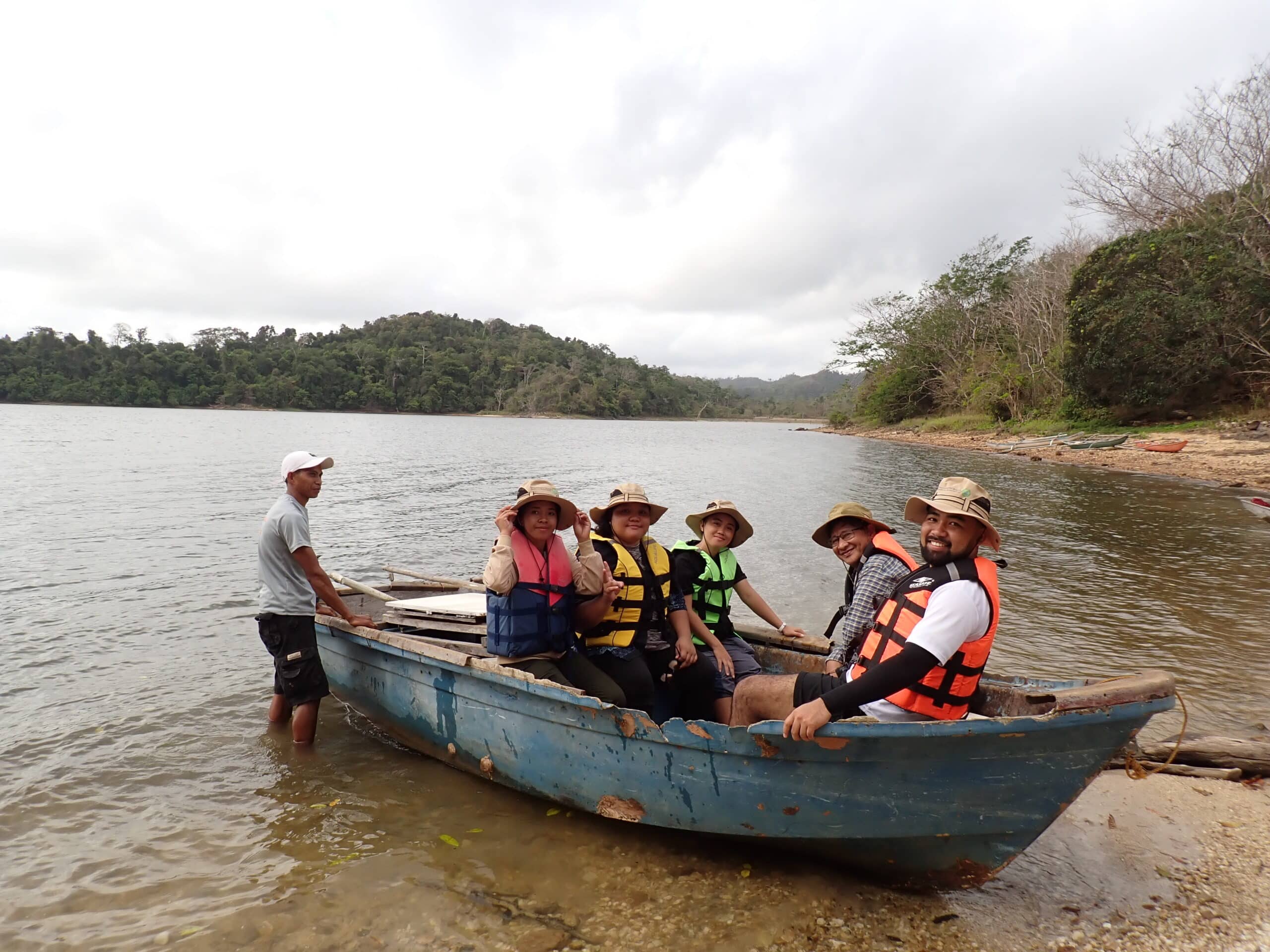 Five people in life jackets sitting in a small wooden boat, and one person in a life jacket standing in the water next to the boat.